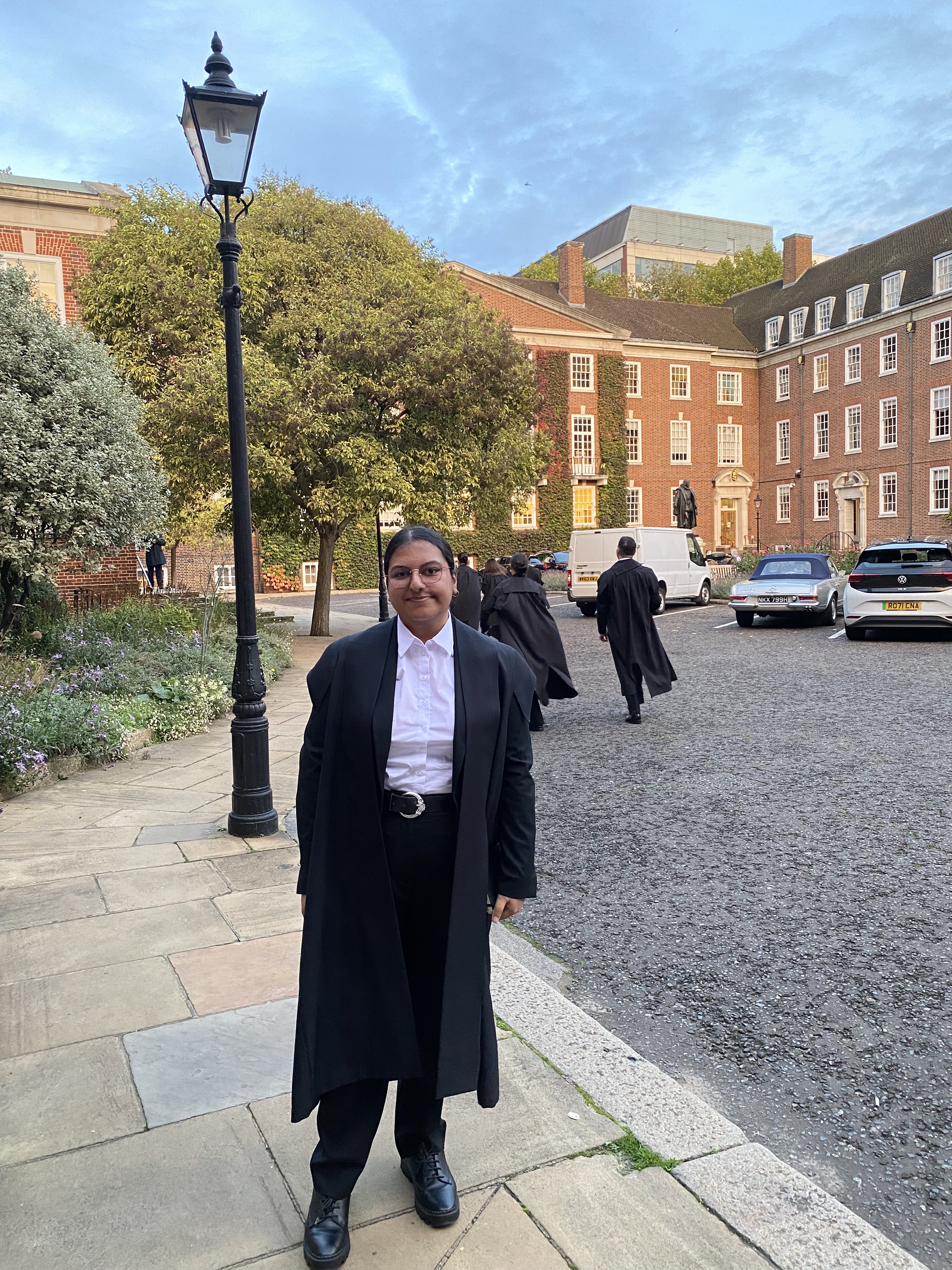 Maria is smiling in a black suit with a white shirt, a black student gown, in front of Gray's Inn.